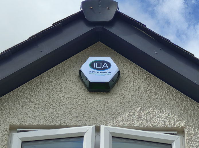 Alarm system sign mounted on the exterior wall of a house, featuring the logo of IDA Alarm Systems Ltd., against a background of blue sky and clouds.