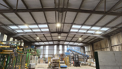 Interior of a large industrial warehouse with a high metal roof, large skylights, and various equipment like stacks of materials, shelving, and a conveyor system. Bright lighting illuminates the space, highlighting its organized layout and extensive storage capacity.