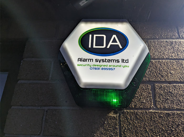 A green-lit hexagonal alarm system sign mounted on a brick wall, featuring the text 