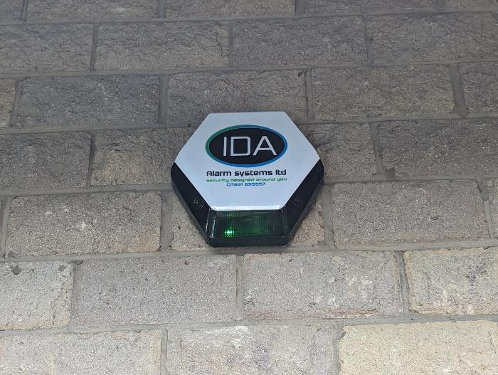 Alarm systems notification sign with a green light mounted on a brick wall, displaying the logo of IDA Alarm Systems Ltd and security information.