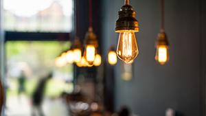 Warm glowing vintage light bulbs hang from the ceiling, creating a cozy atmosphere in a café setting, with blurred figures in the background.