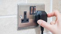 A hand is plugging a black electrical plug into a metallic wall socket with multiple outlets.