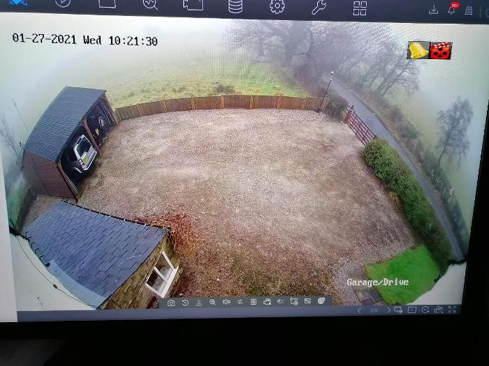 Aerial view of a foggy driveway and garage area with gravel surface and wooden fence, showing a parked car in the garage and a gated road entrance. Ideal for property security monitoring.